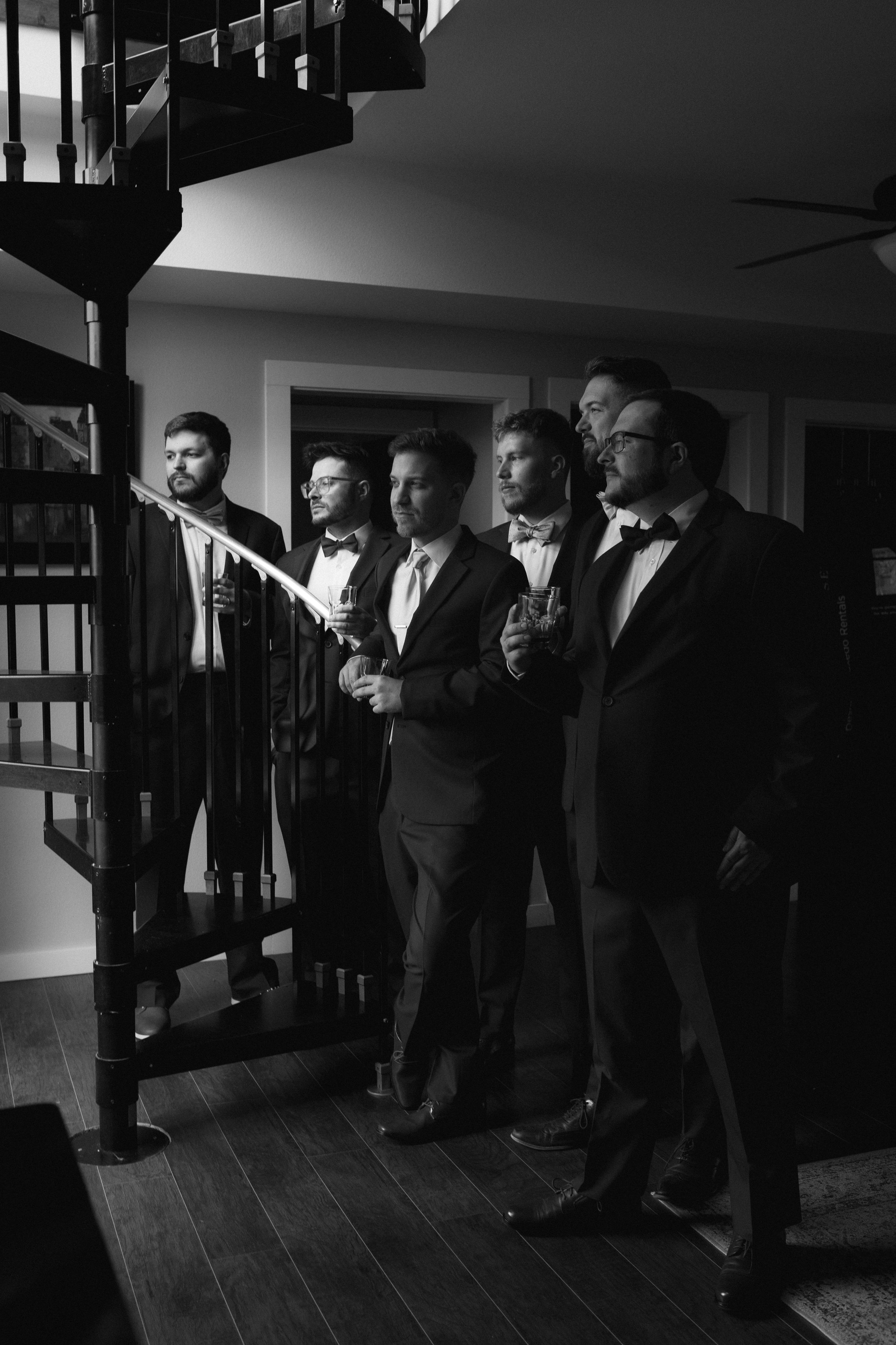 Groomsmen line up in dark suits against dramatic lighting in black and white photo.