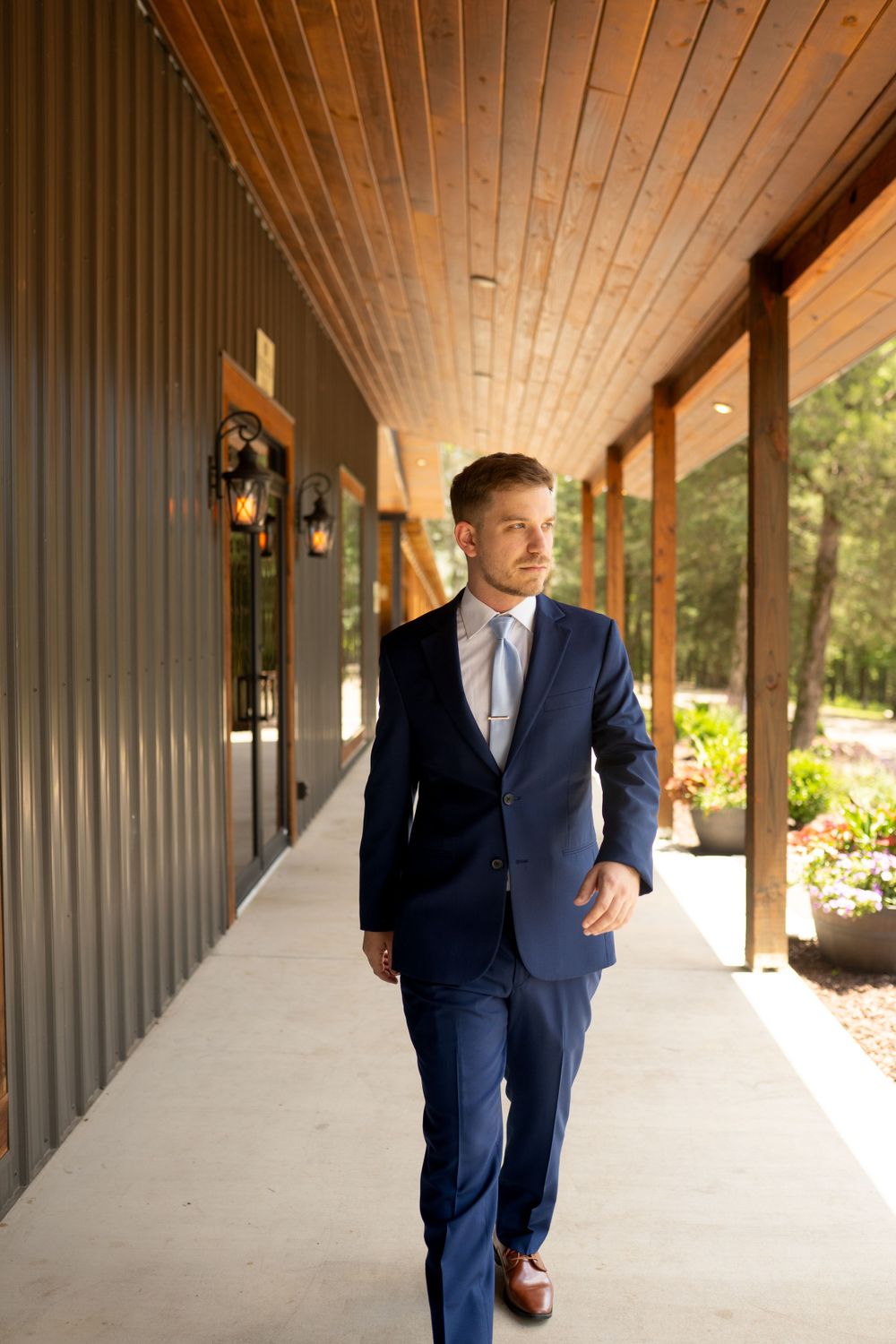 Groom in navy suit runs playfully down covered walkway with wooden ceiling.