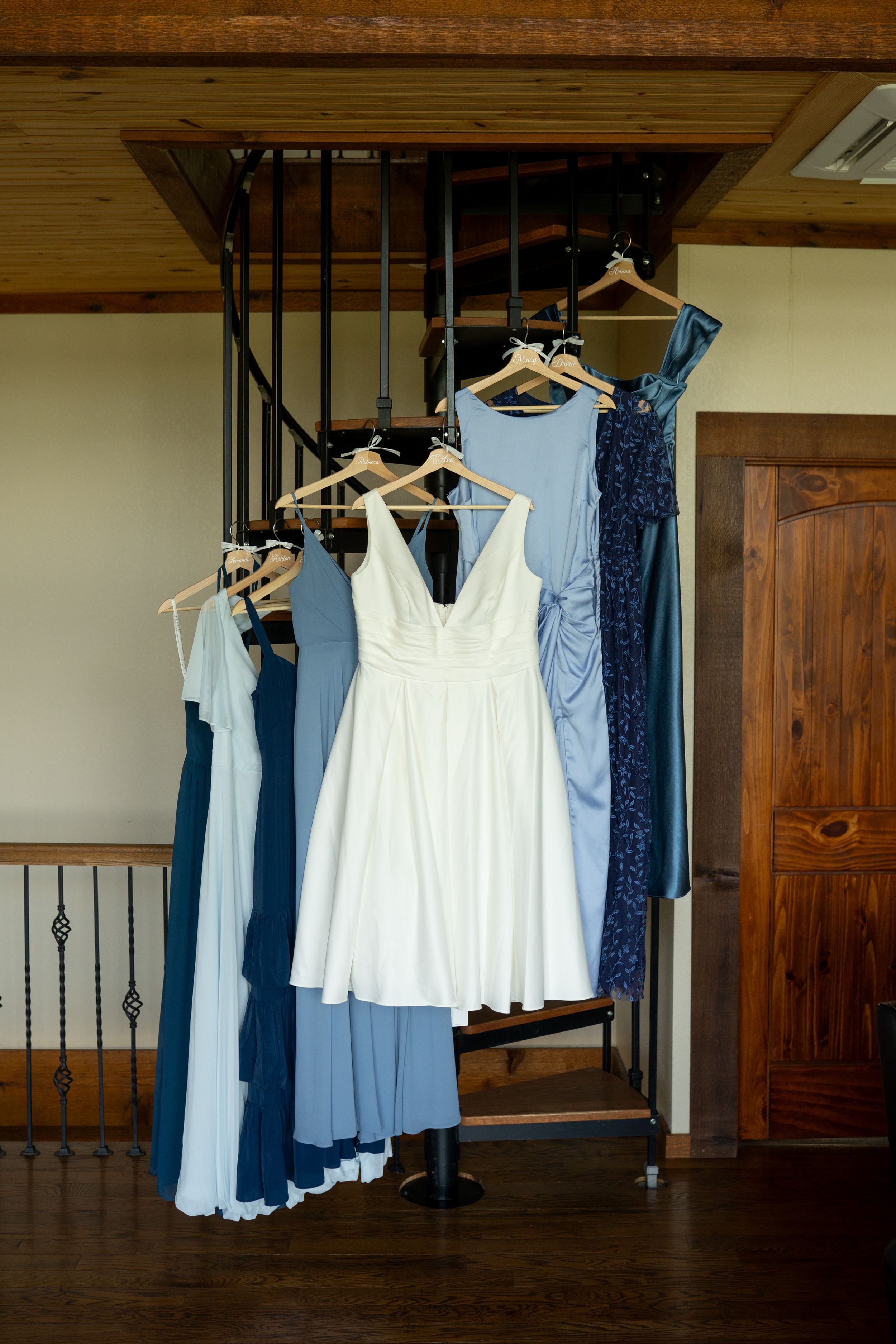 Wedding dresses and bridesmaid gowns hanging on rack against wooden wall.