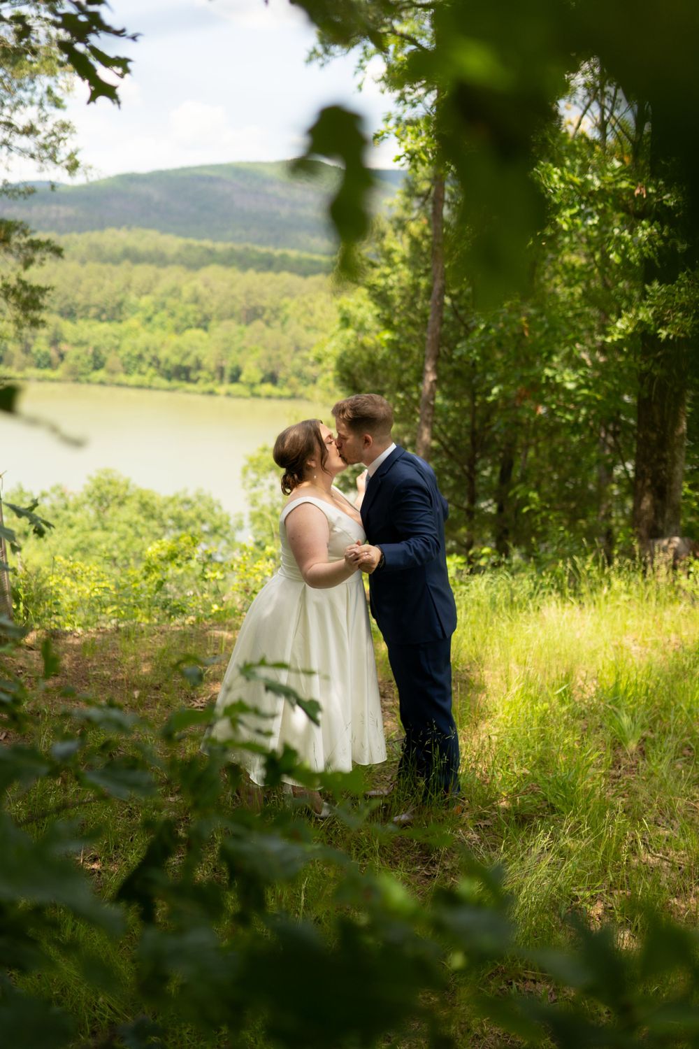 Wedding couple poses in a scenic hillside meadow with mountains and greenery in the background.