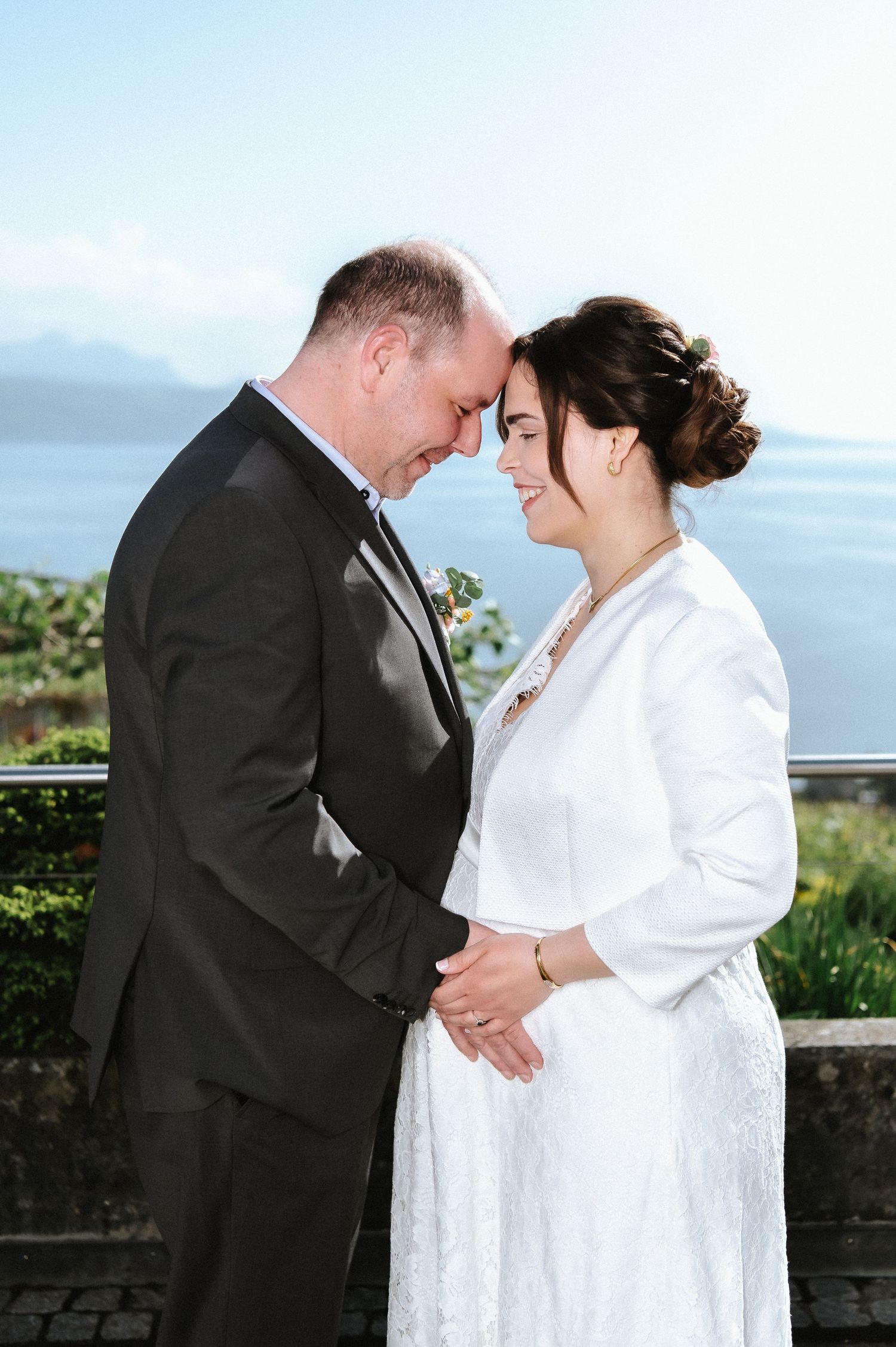 A couple in wedding attire embraces on a scenic overlook with water views.