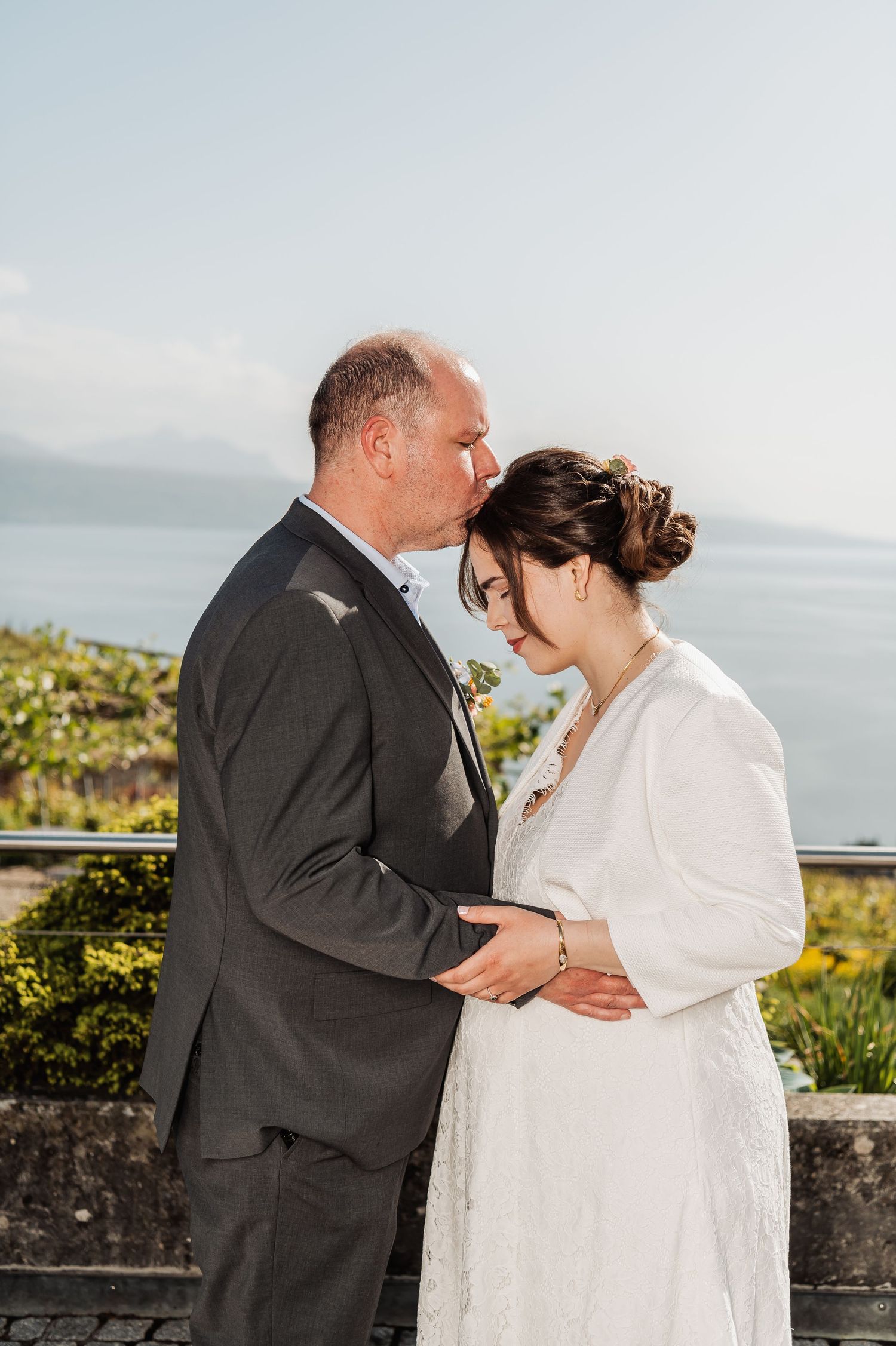 A romantic wedding couple embraces against a scenic coastal backdrop with lush greenery and ocean views.