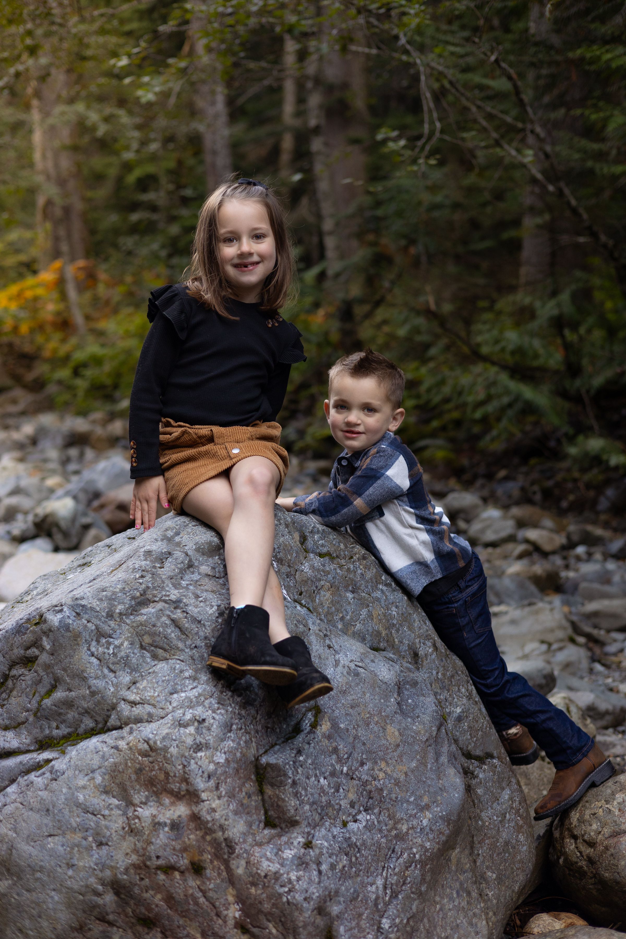 Brother and sister sit on a large river rock near Snoqualmie and smile at photographer Jess Bickel during their family session.