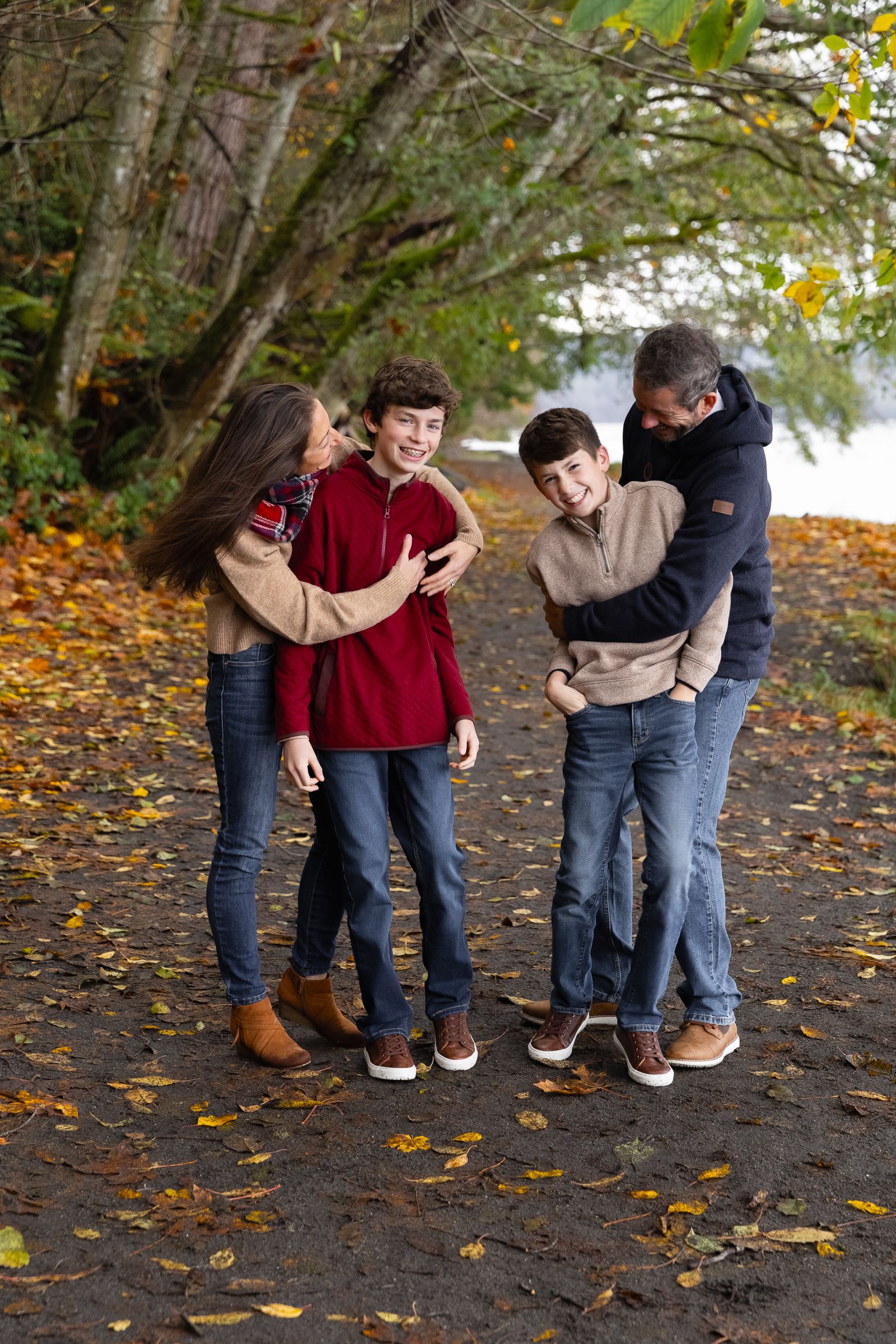 Mom and dad hug their tween sons under large trees in autumn at Owen Beach in Tacoma, Washington.