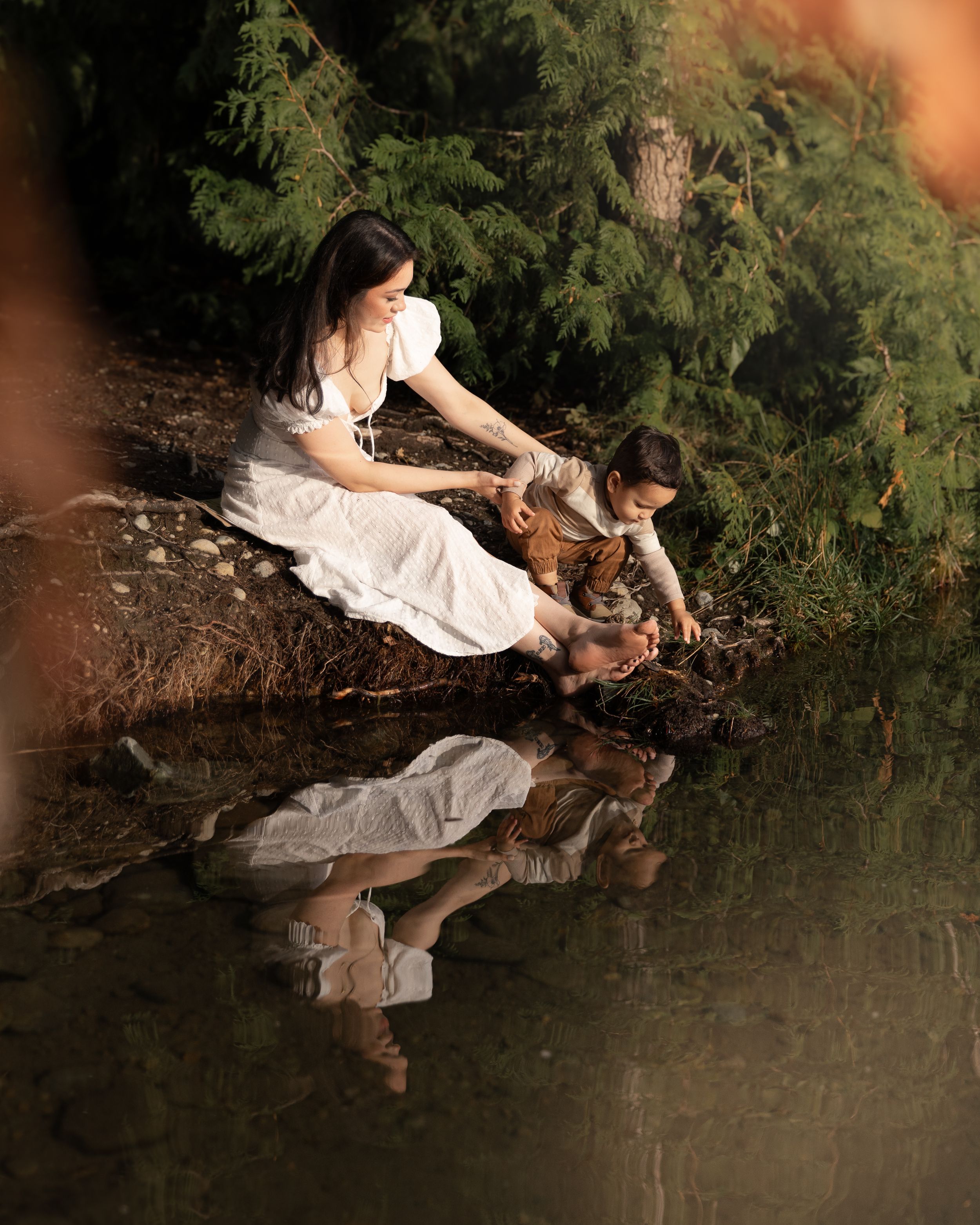 A young mom holds her toddler on a bank at Lake Tapps as he reaches for the water. Their reflection is visible in the water.