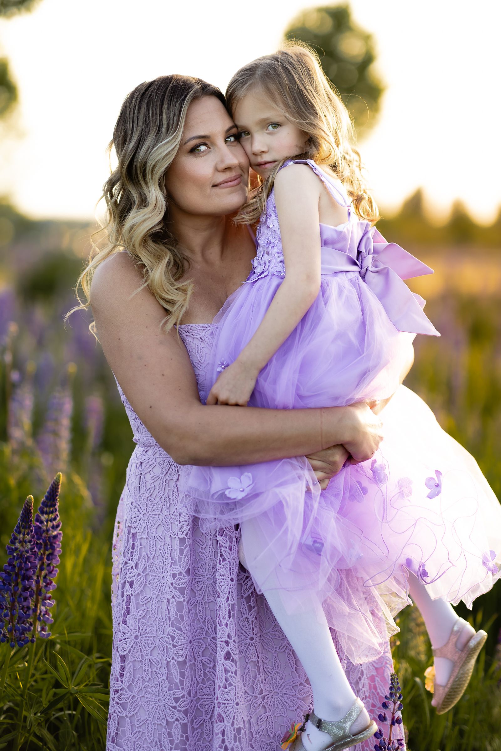 Mom holds her daughter in a field of purple wildflowers. They are backlit by the setting sun during a family session near Seattle.