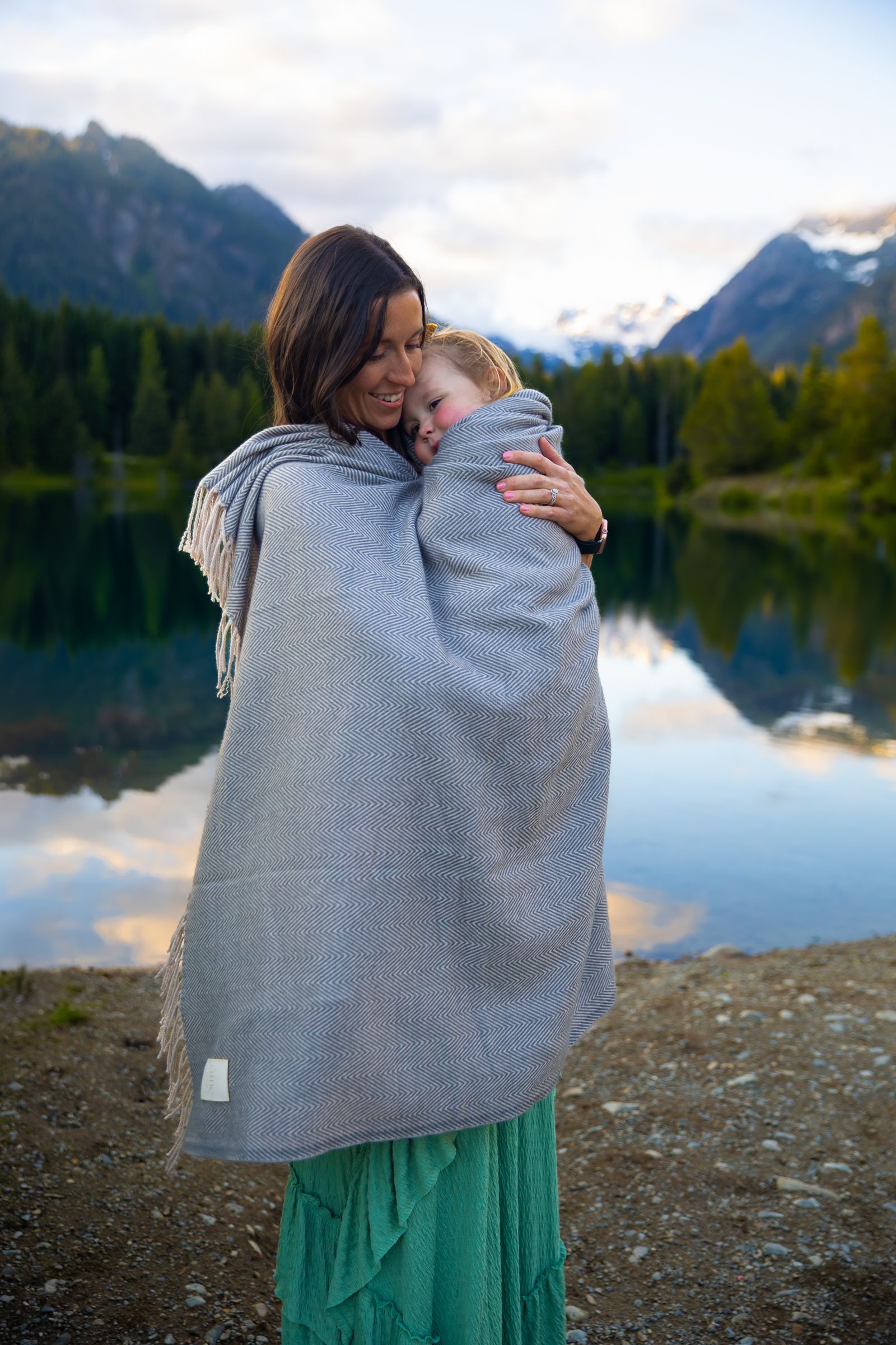 Mom wrapped up in blanket with young daughter in front of lake and mountain landscape in Washington.