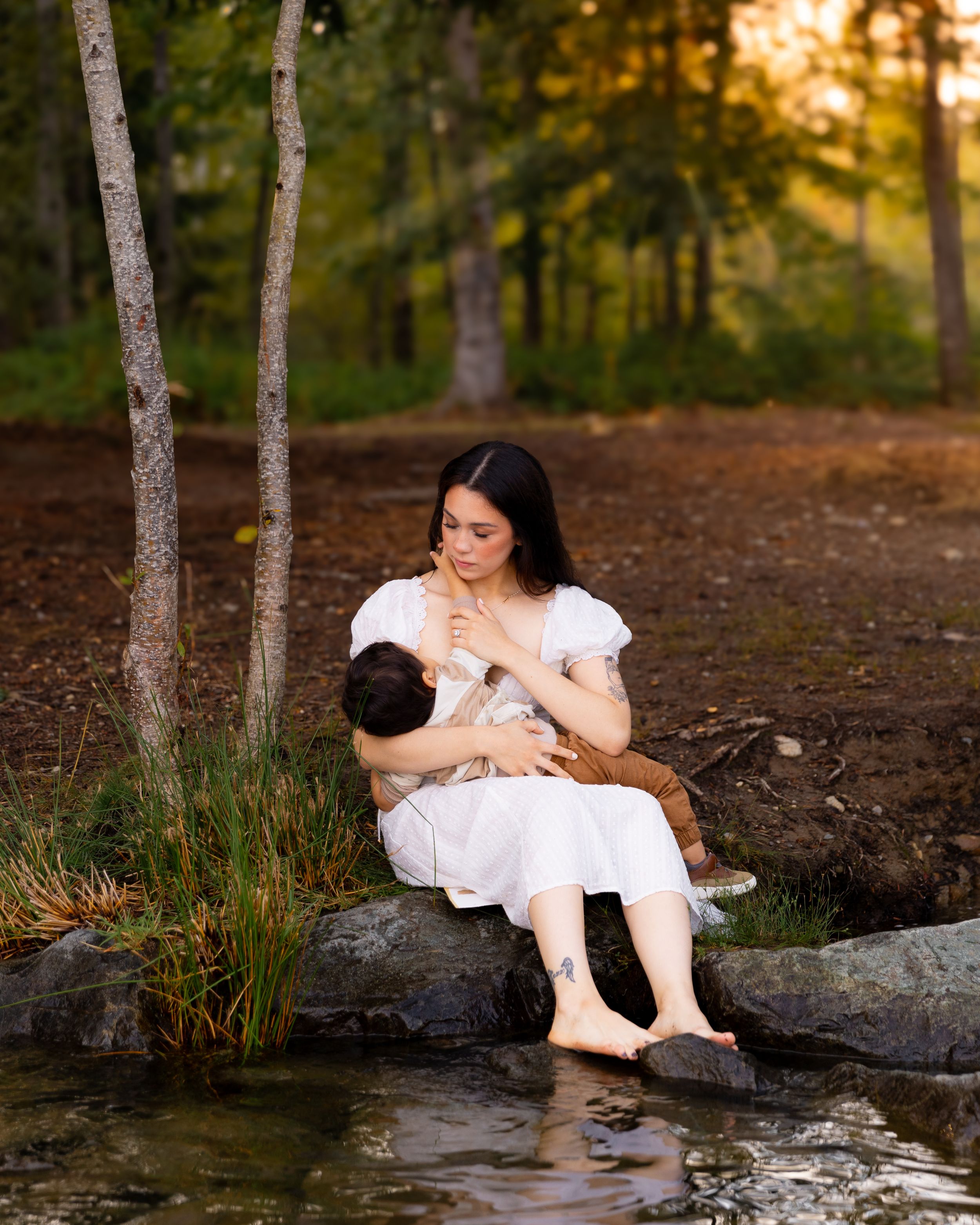 A young mom nurses her toddler at sunset in a forested area with her feet dangling over Lake Tapps in Washington.