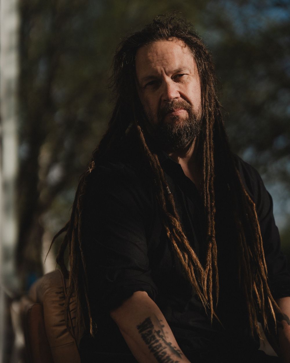A person with long dreadlocks stands outdoors wearing dark clothing in natural lighting.