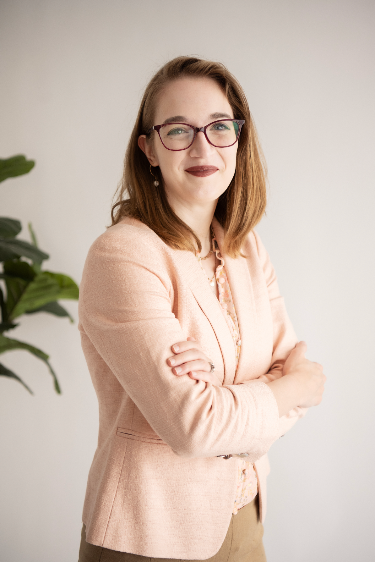 Professional portrait of person in peach blazer and glasses with arms crossed against light background.