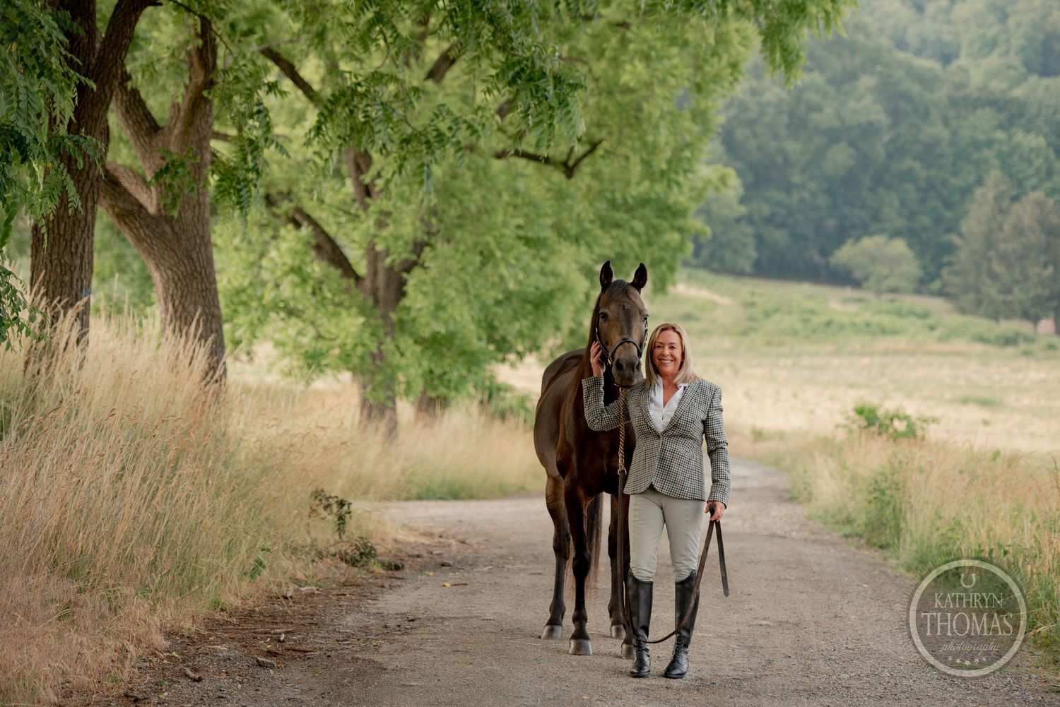 Person in business attire walks alongside a bay horse on a peaceful wooded trail.