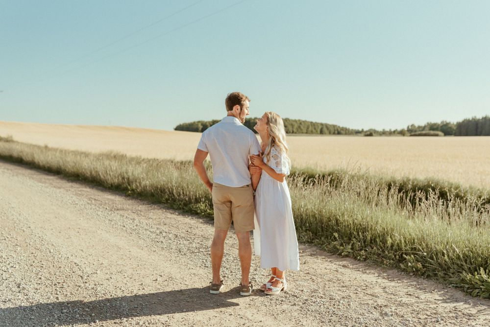 Couple standing together on dirt road surrounded by golden wheat fields at sunset.