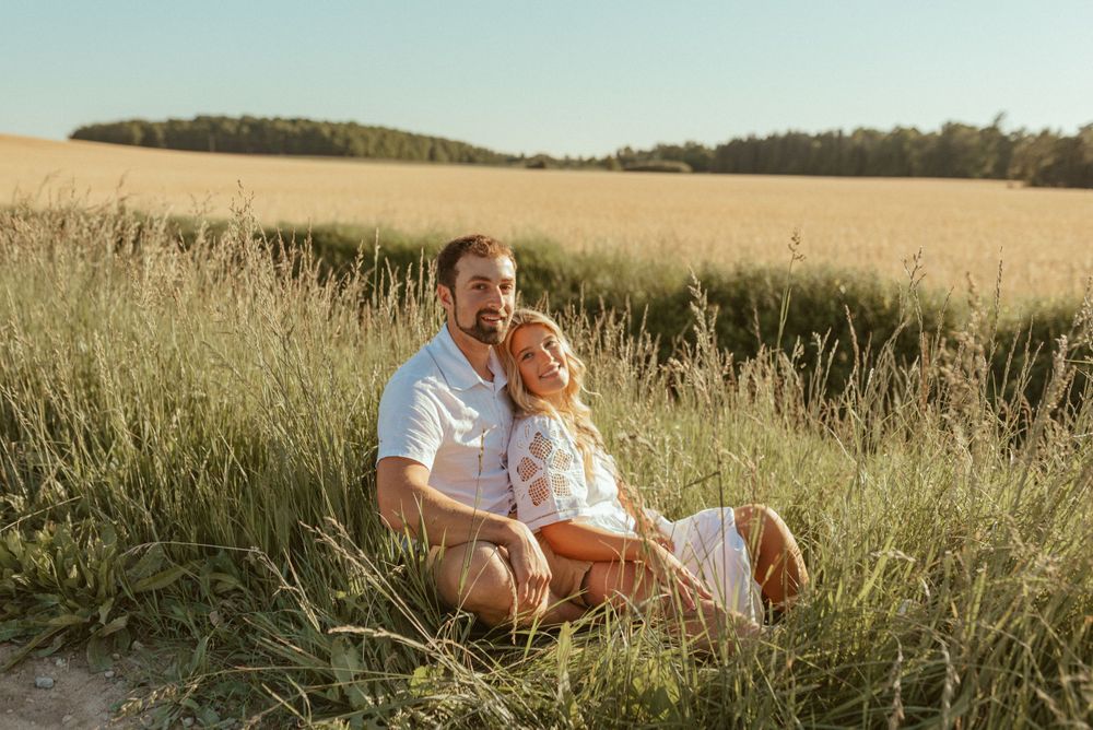 Couple embracing while sitting in sunlit grass field with rolling hills in background.