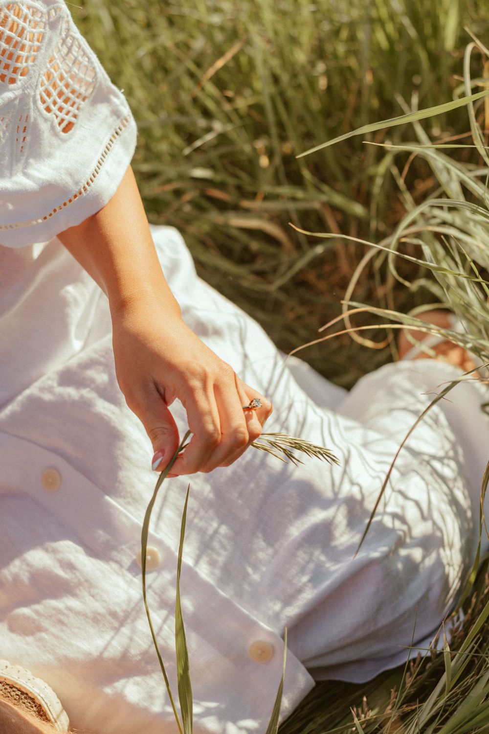 Close-up of hand touching tall grass while wearing white lace dress in sunlit field.