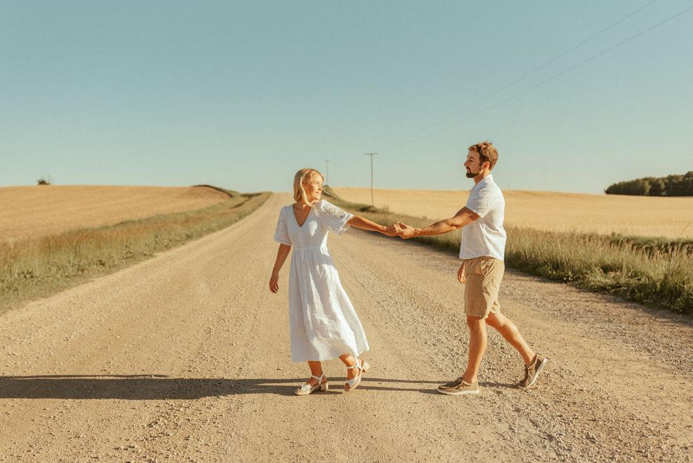Couple dancing on dirt road surrounded by golden wheat fields under blue sky.