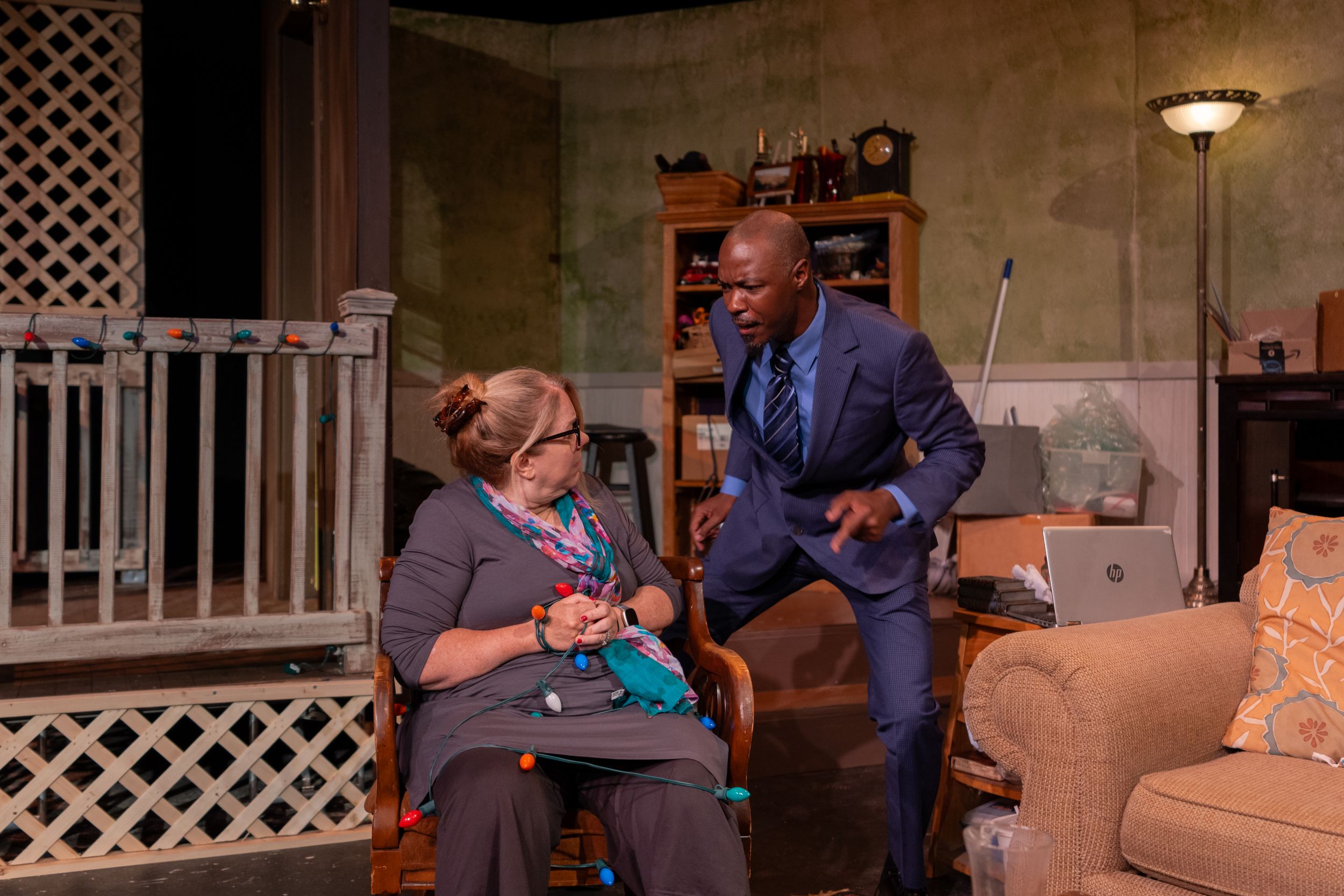 A dramatic scene unfolds between two performers in a theatrical living room.