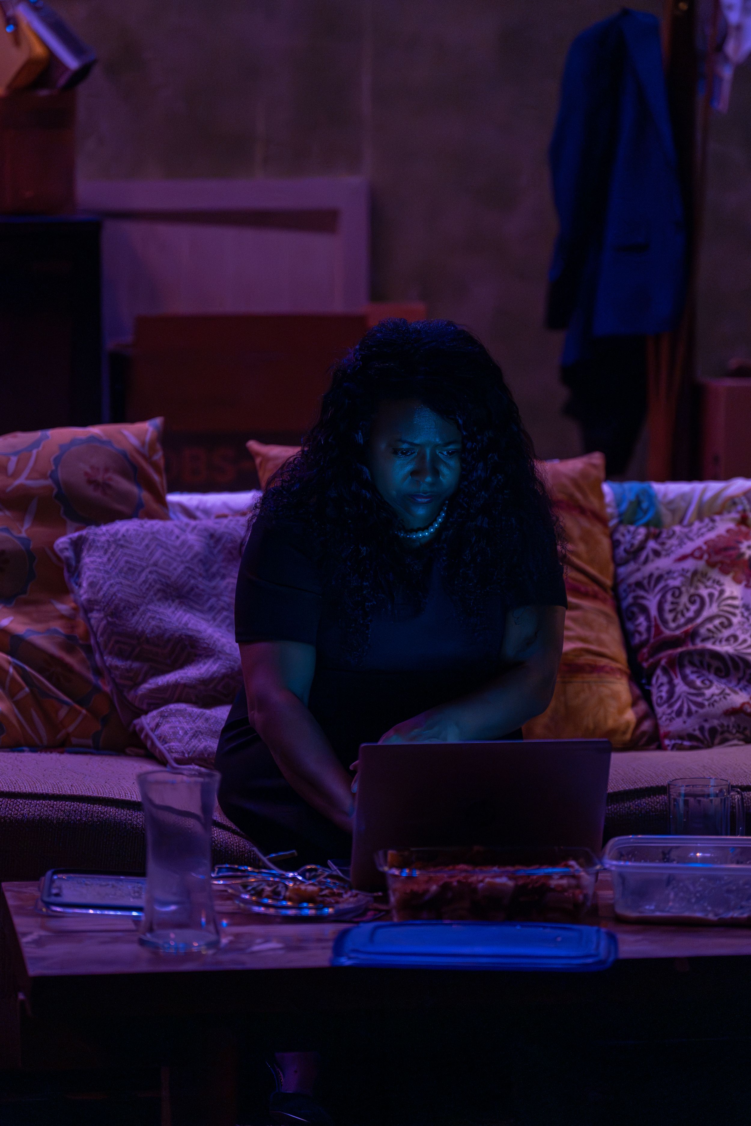 A person sits on a couch with a laptop in moody purple lighting.