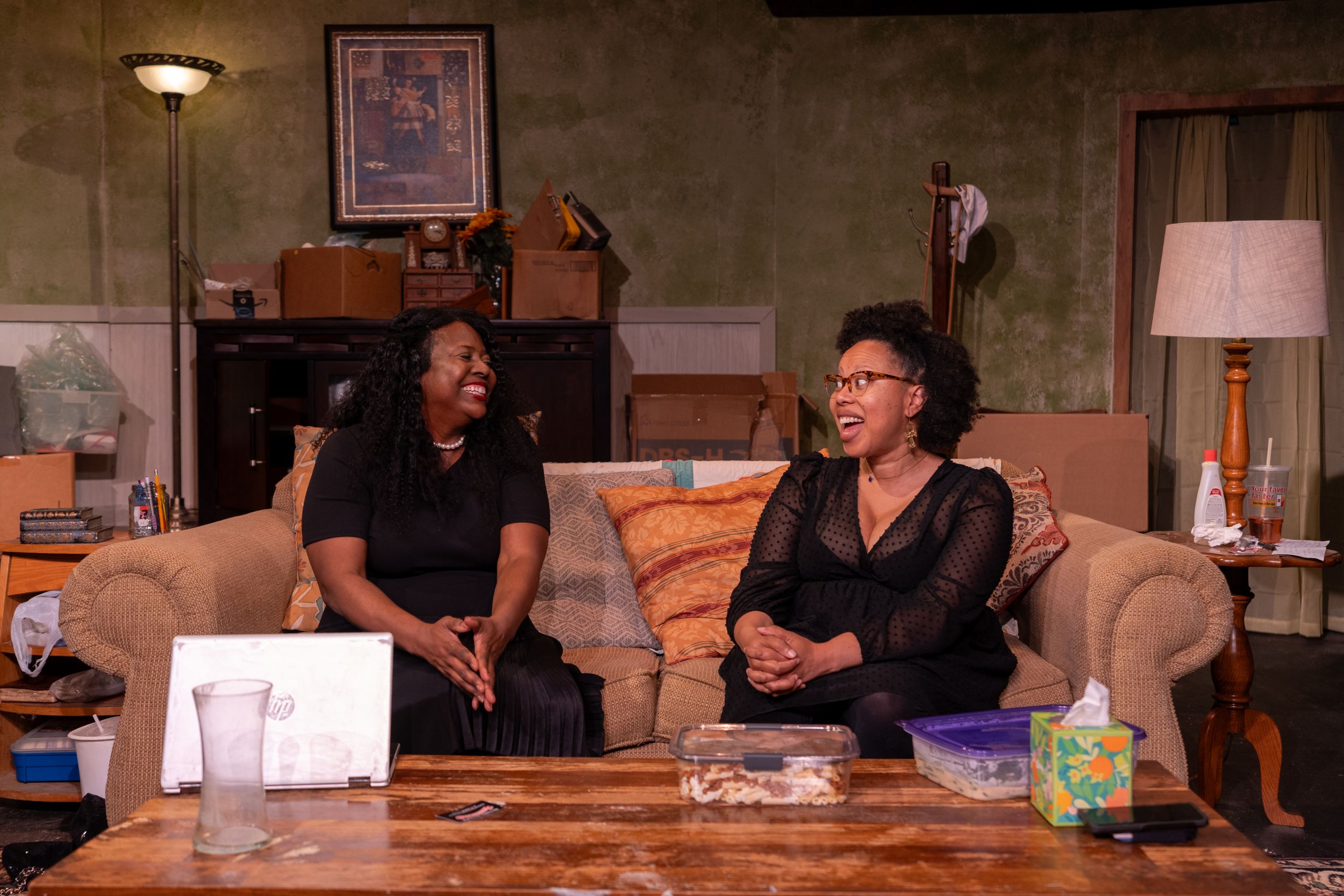 Two people share a conversation while sitting on a couch in a dimly lit living room setting.