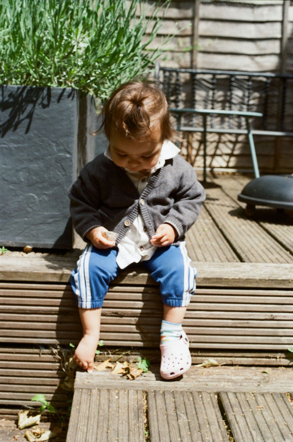 Toddler in gray cardigan and blue pants sits on wooden deck steps, looking down at something in hands.