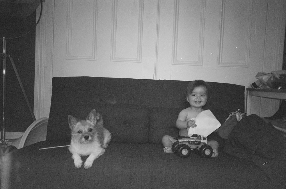 Black and white photo of a smiling baby sitting on a couch with a toy truck beside a fluffy dog in a living room.