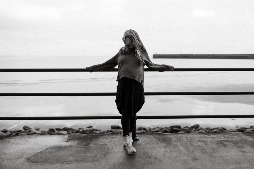 A blonde woman leans against a metal railing overlooking a calm sea in a black and white outdoor photo.