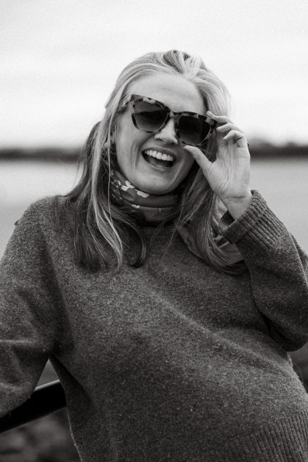 A laughing blonde woman in sunglasses and a gray sweater poses outdoors near water in a black and white photo.