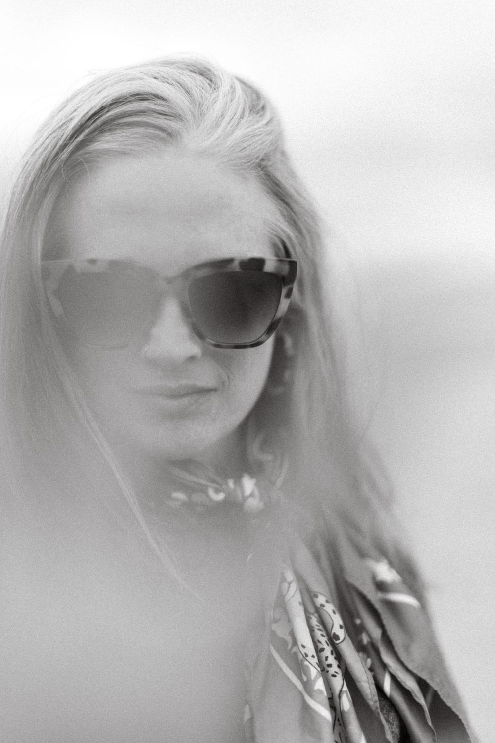 Black and white portrait of a woman with long hair wearing large sunglasses and a patterned scarf outdoors.