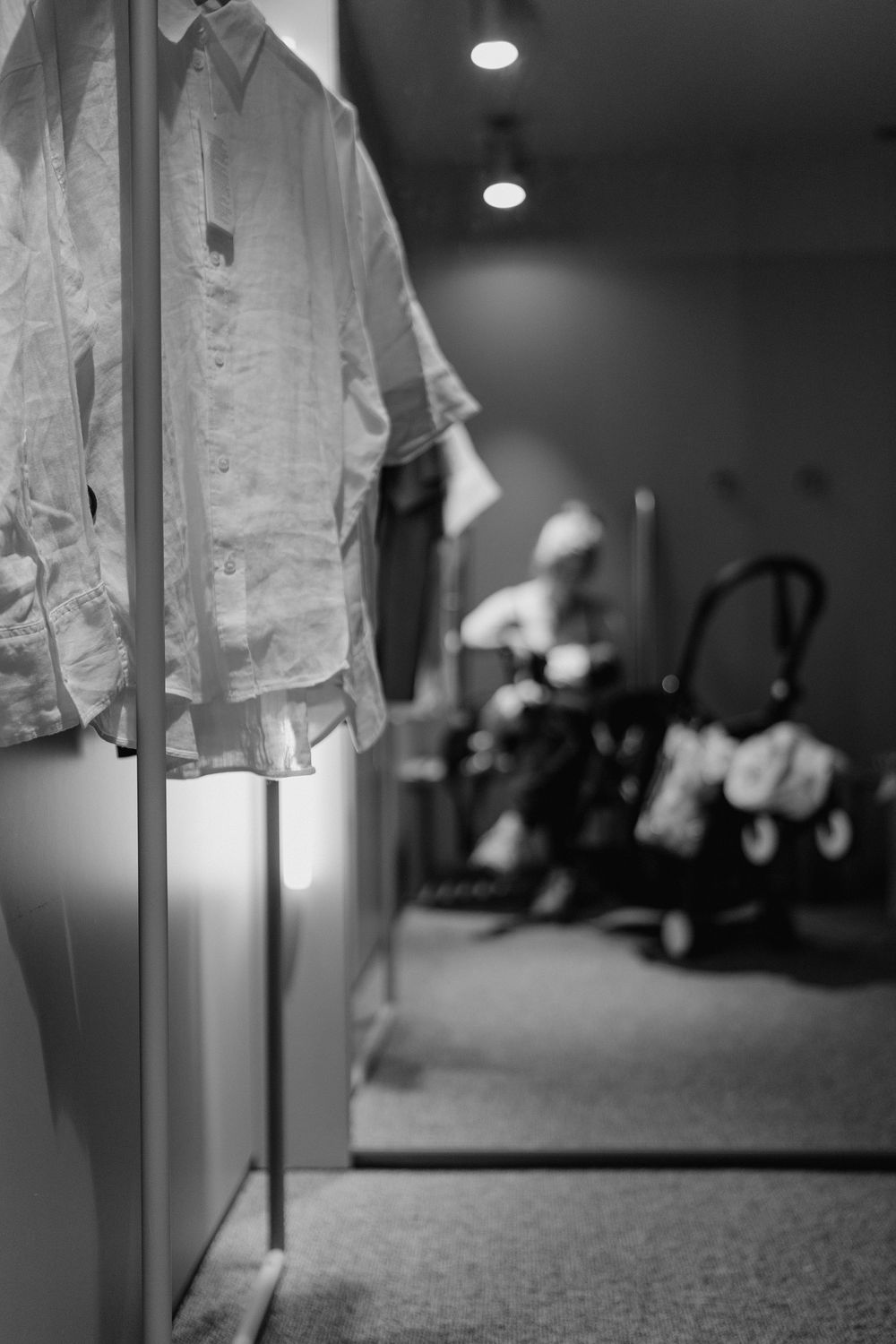 Black and white photo of a white shirt hanging in a dressing room with a blurred figure and stroller in background.