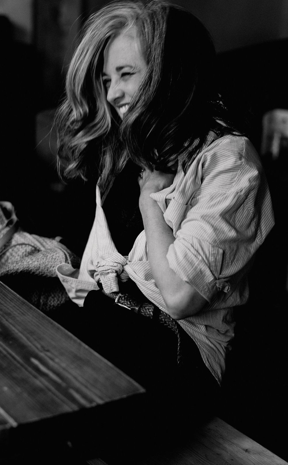 Black and white photo of a smiling woman with wavy hair leaning over a wooden table indoors.