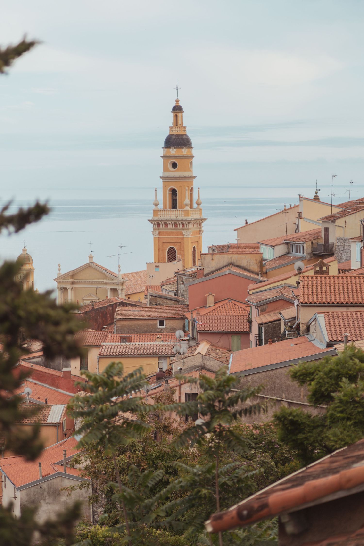 Scenic view of terracotta rooftops and historic church bell tower in Mediterranean coastal town against cloudy sky.