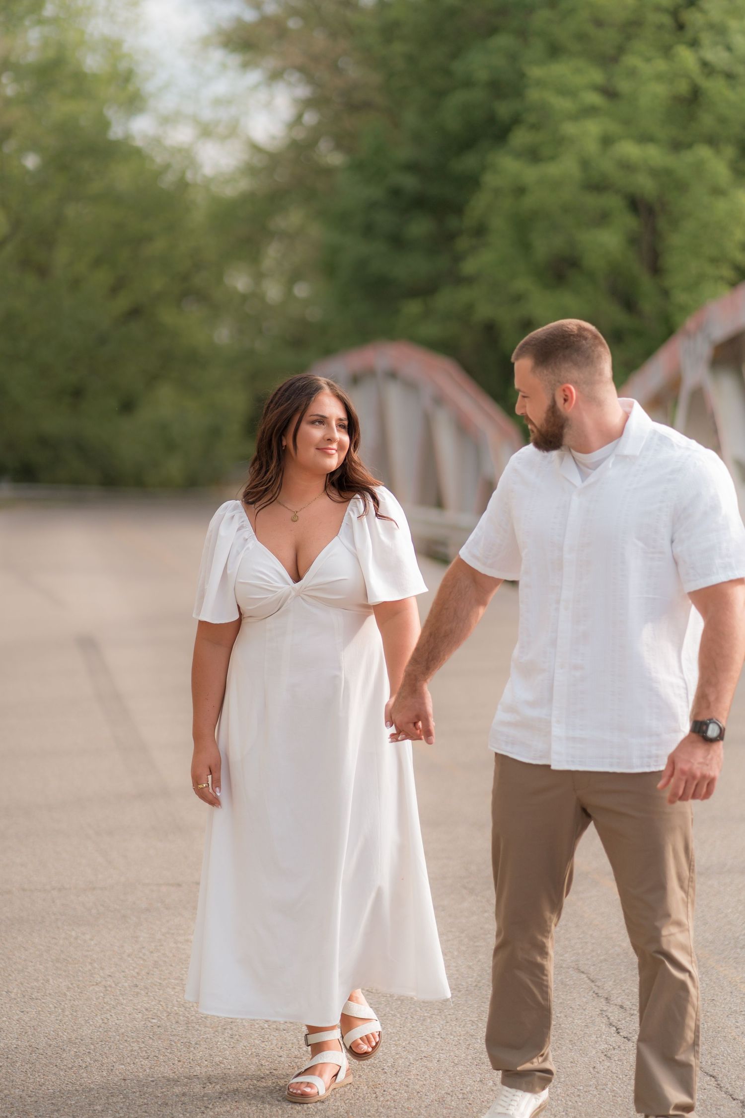 A romantic couple in white outfits walking hand in hand on a path with a bridge in the background.