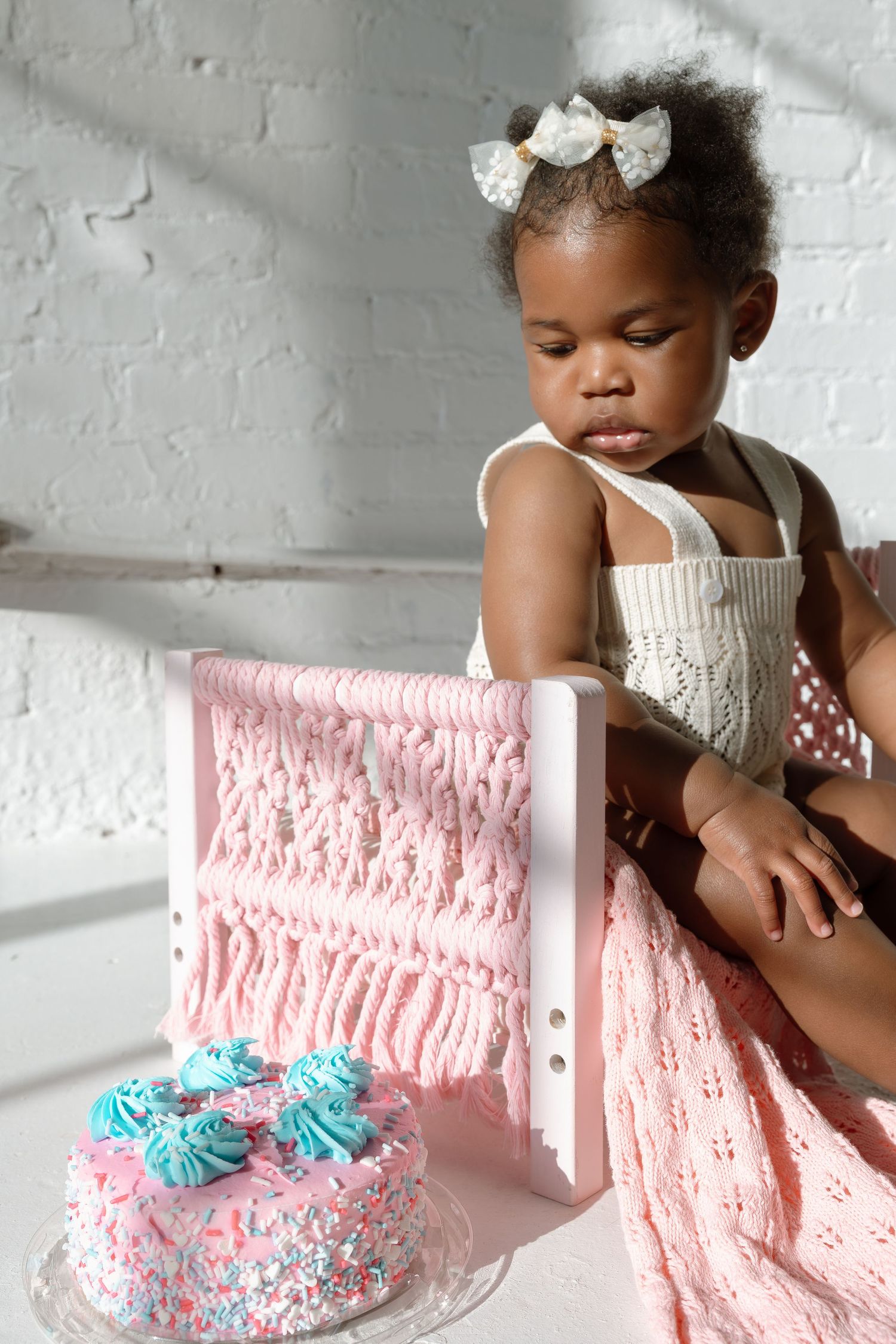 A small child in a white dress sits beside a pink and blue decorated birthday cake on a soft pink backdrop.