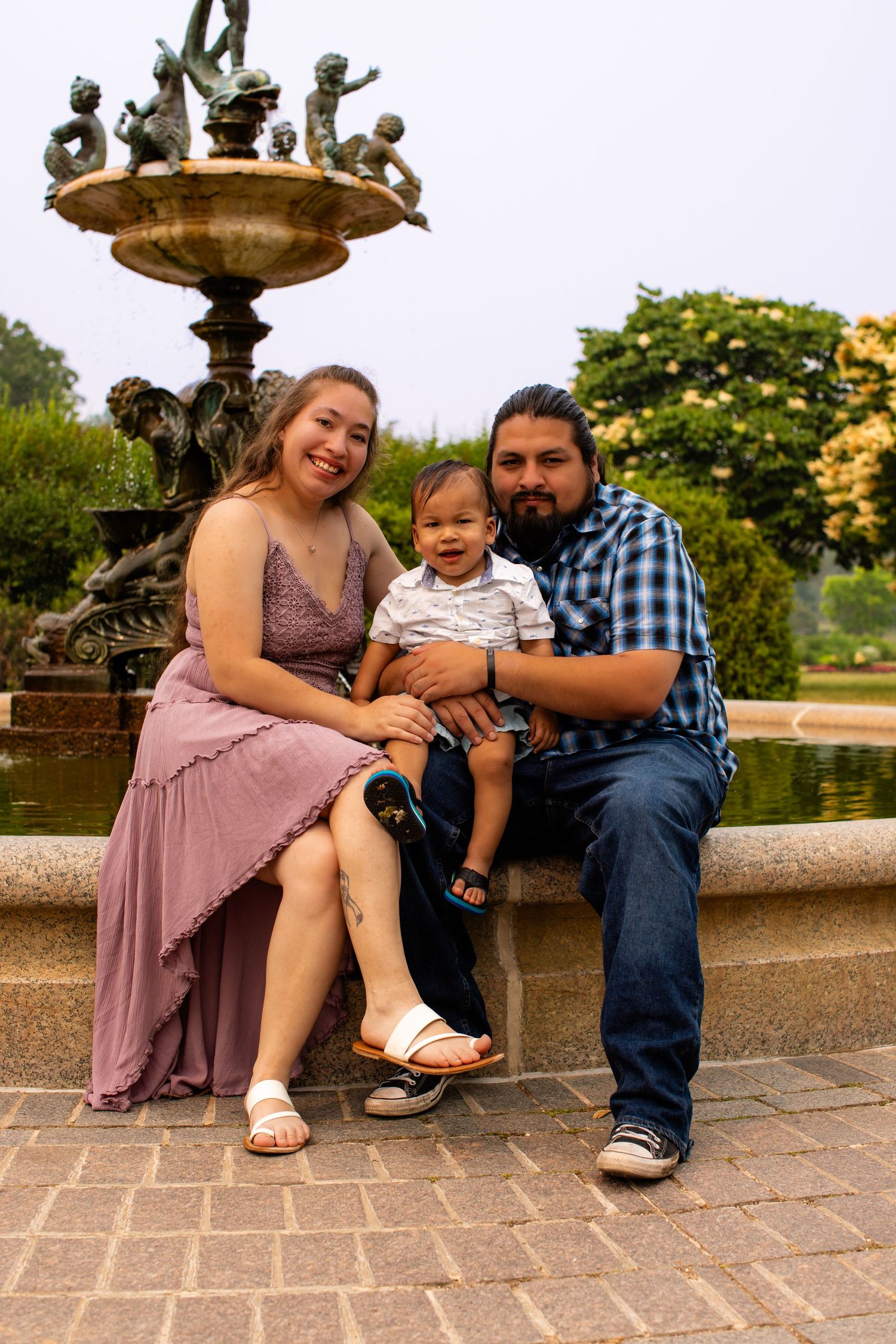 A family poses together on a brick ledge in front of an ornate fountain in an outdoor garden setting.