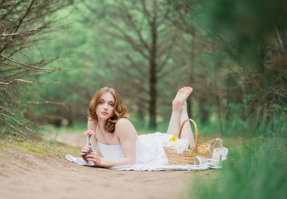 Young woman in white dress lying on blanket in pine forest holding flower near wicker picnic basket.