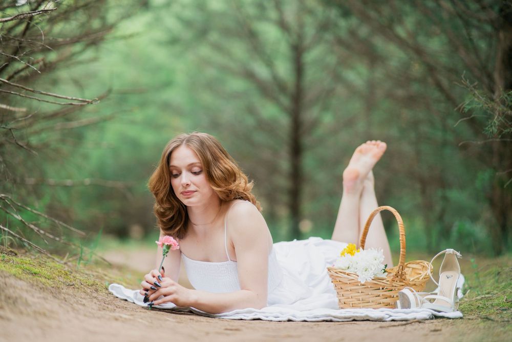 Young woman in white dress lying on blanket writing in pine forest beside wicker picnic basket.