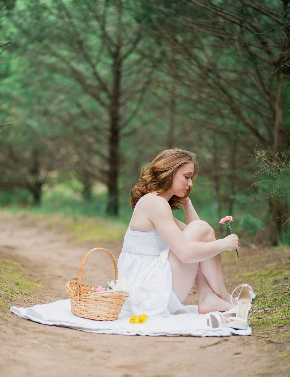 Young woman in white dress sits on blanket in forest, holding a pink flower beside a wicker picnic basket.