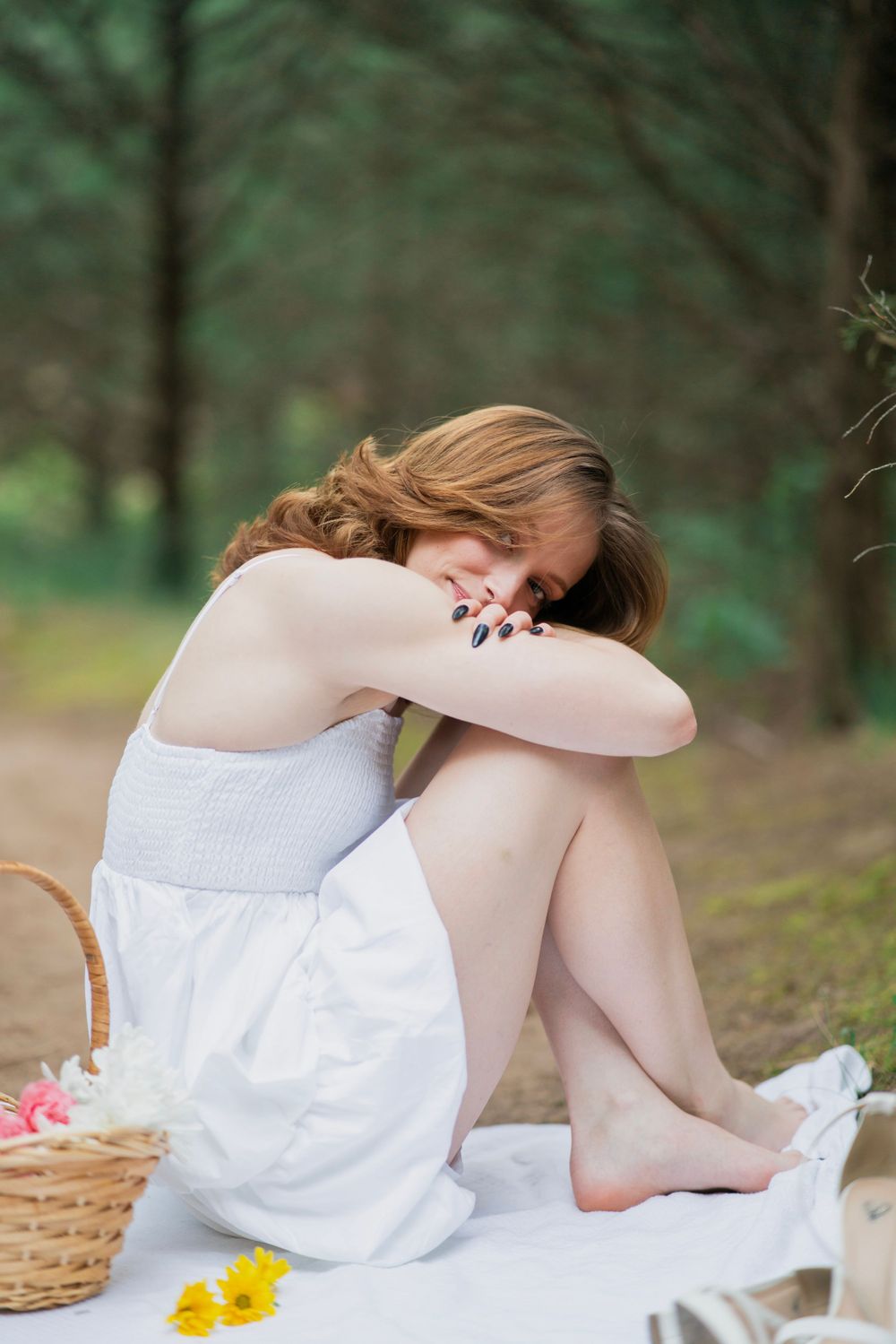 Young woman in white sundress sits barefoot on blanket in forest, hugging her knees with head down.