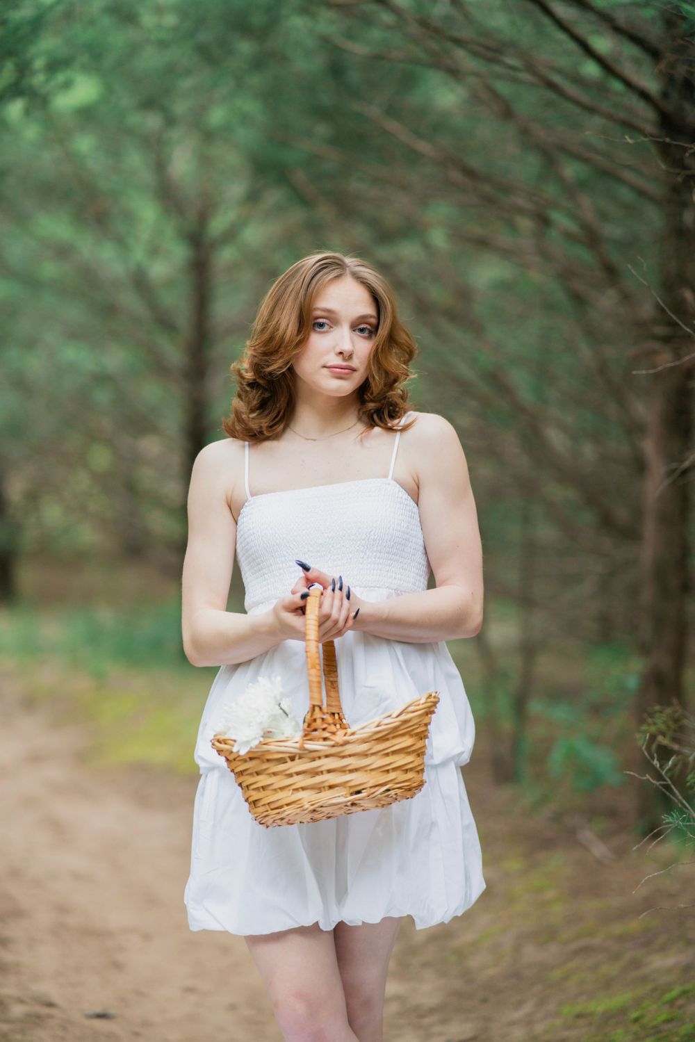 Young woman in white sundress walks forest path carrying a wicker basket, looking at camera.