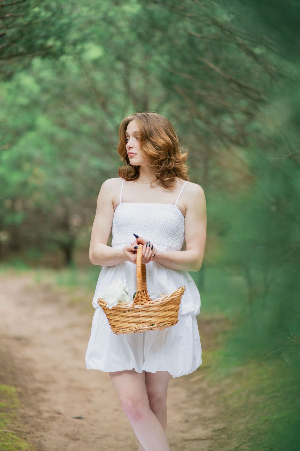 Young woman in white dress holds wicker basket while standing on a sandy forest path among green trees.