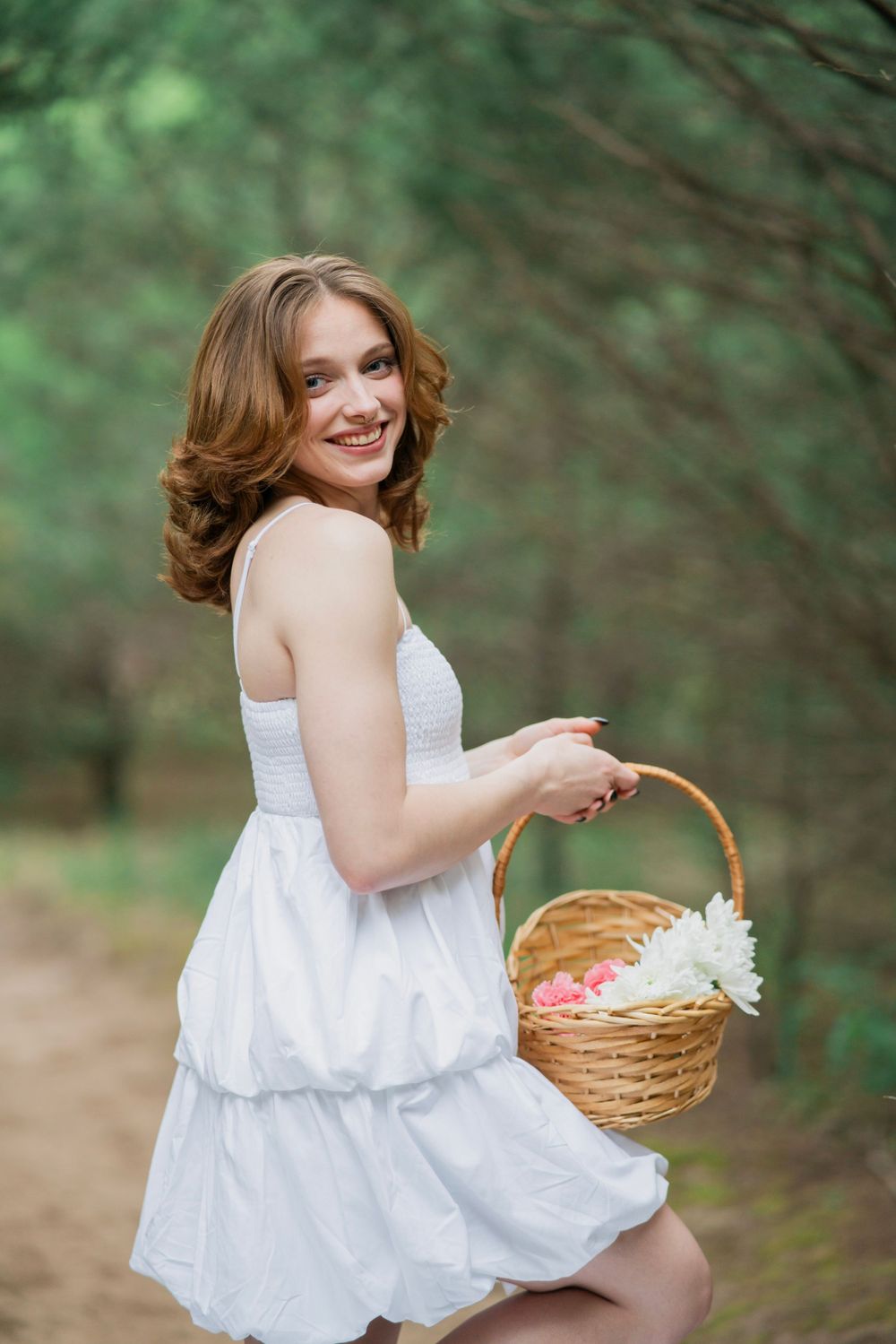 Smiling young woman in white dress poses on forest path holding a wicker basket filled with white flowers.