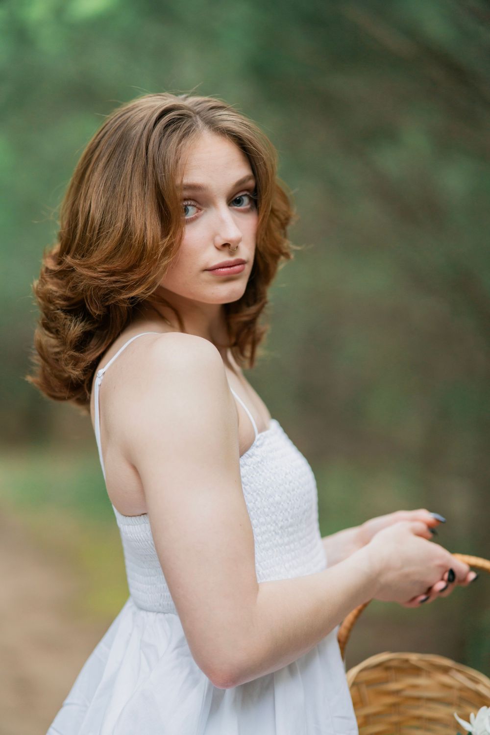 Young woman in white dress looks over shoulder while holding a wicker basket on a shaded forest path.