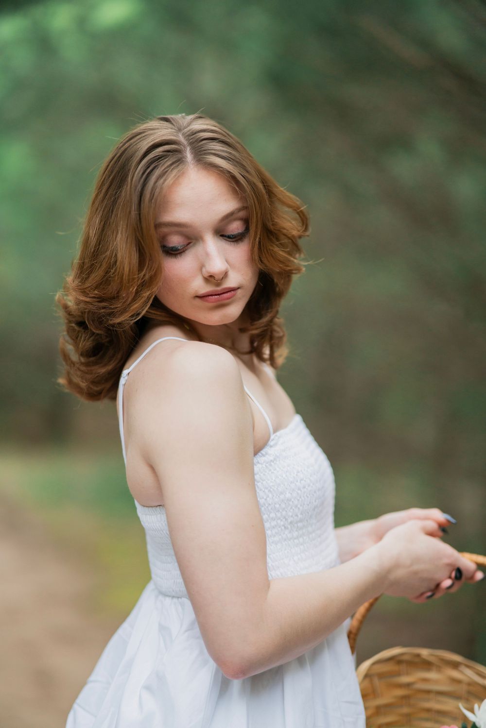 Young woman in white sundress looks down while holding a wicker basket on a green forest path.