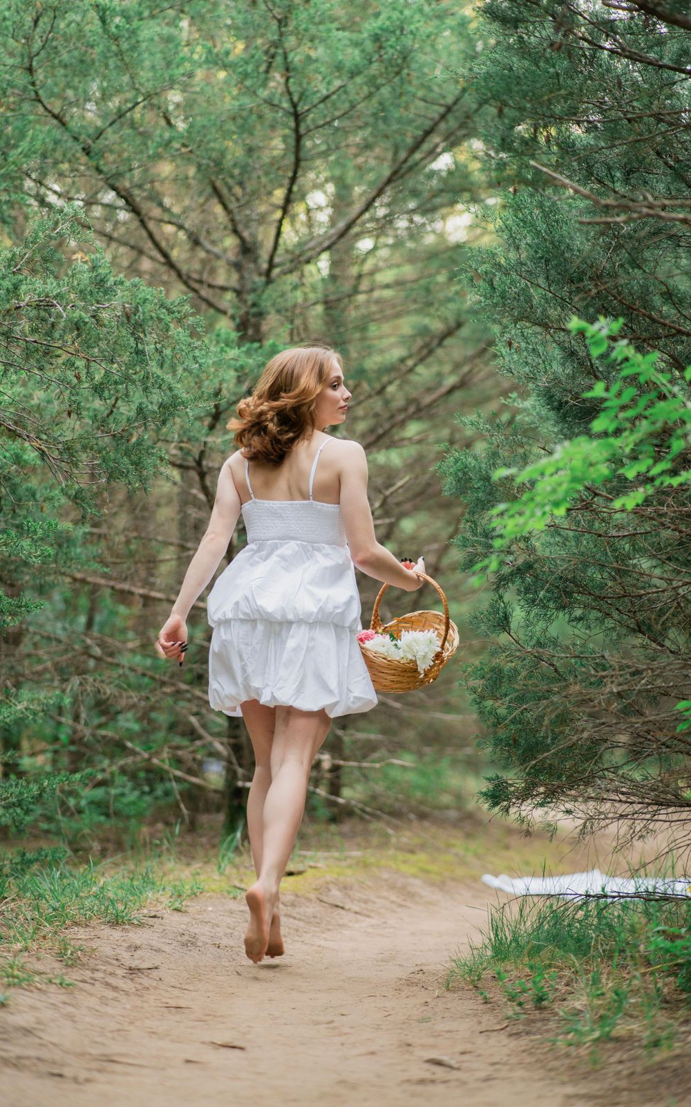 Young woman in white dress walks barefoot away down a sandy forest path carrying a flower-filled wicker basket.