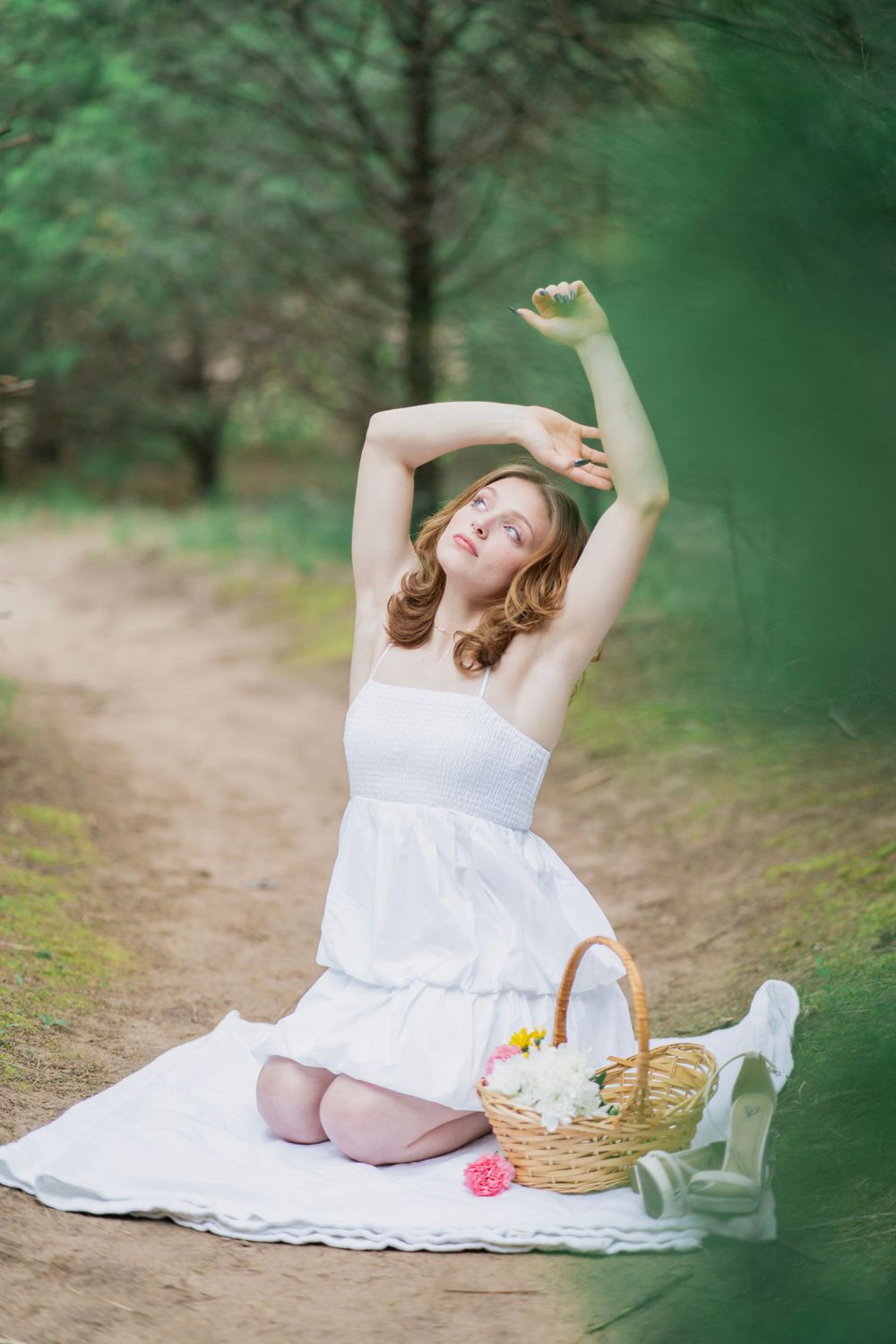 Dreamy young woman in white dress kneeling with arms raised on forest path beside wicker picnic basket.