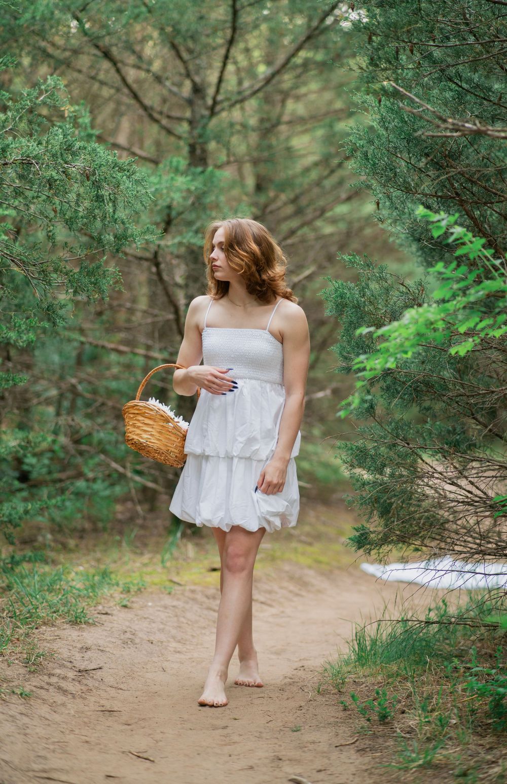 Young woman in white ruffled sundress holds wicker basket while walking barefoot on woodland trail.