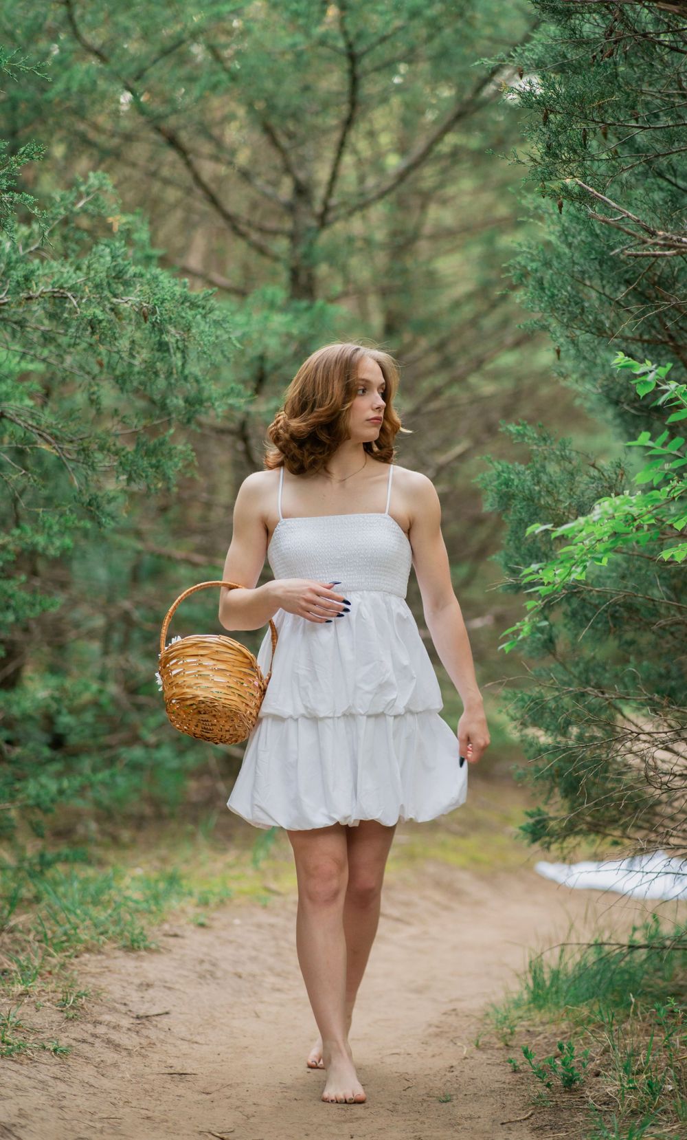 Barefoot woman in white tiered sundress carrying wicker basket walks along sandy forest path.