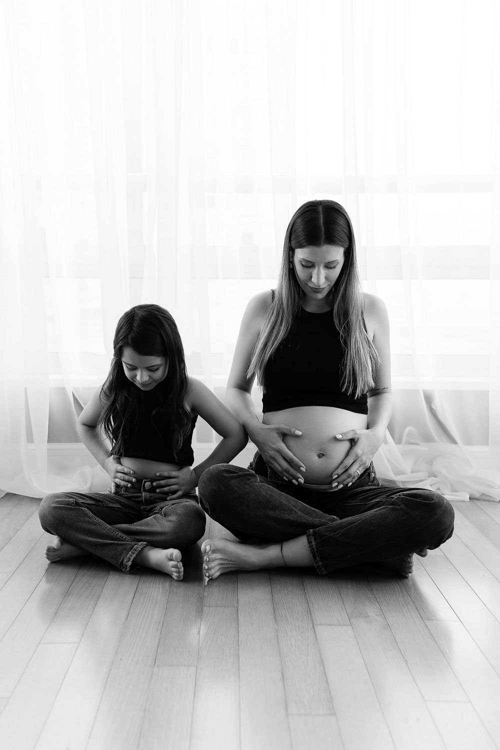 Black and white portrait of mother and daughter touching their bellies in sitting pose.