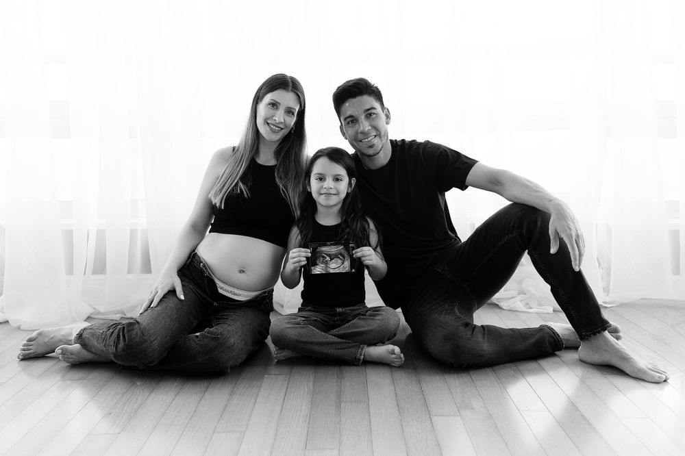 Black and white family maternity photo with mother showing baby bump while sitting on wooden floor.