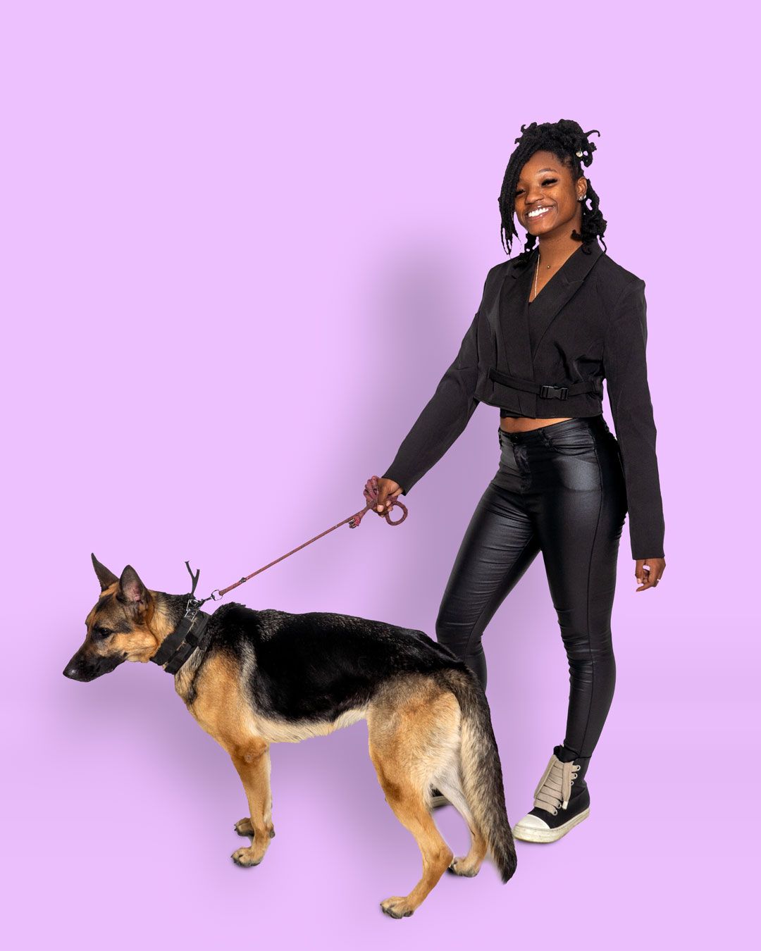 Studio portrait of someone in a black outfit with crop top and leather pants walking a German Shepherd mix dog on pink background.