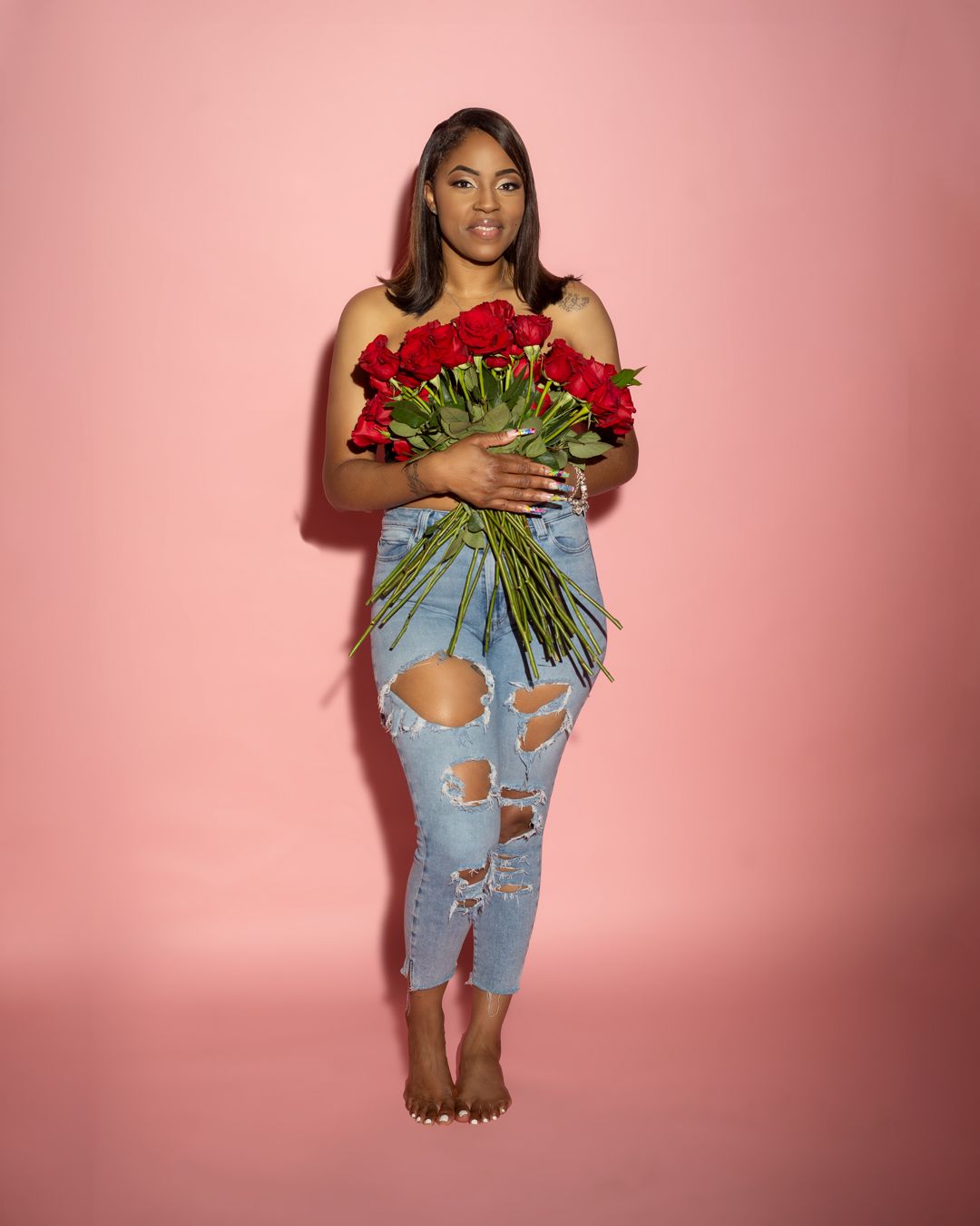 Model in distressed jeans holds a large bouquet of red roses against pink backdrop.